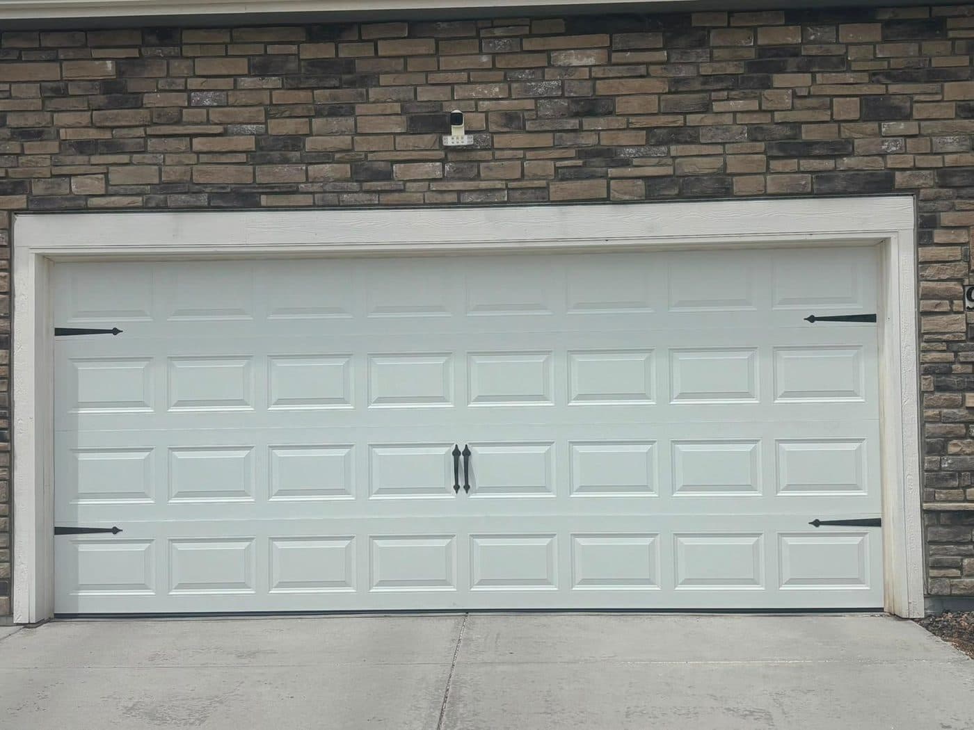 A finished white raised-panel double garage door on a stone-fronted house, with black decorative hinges.