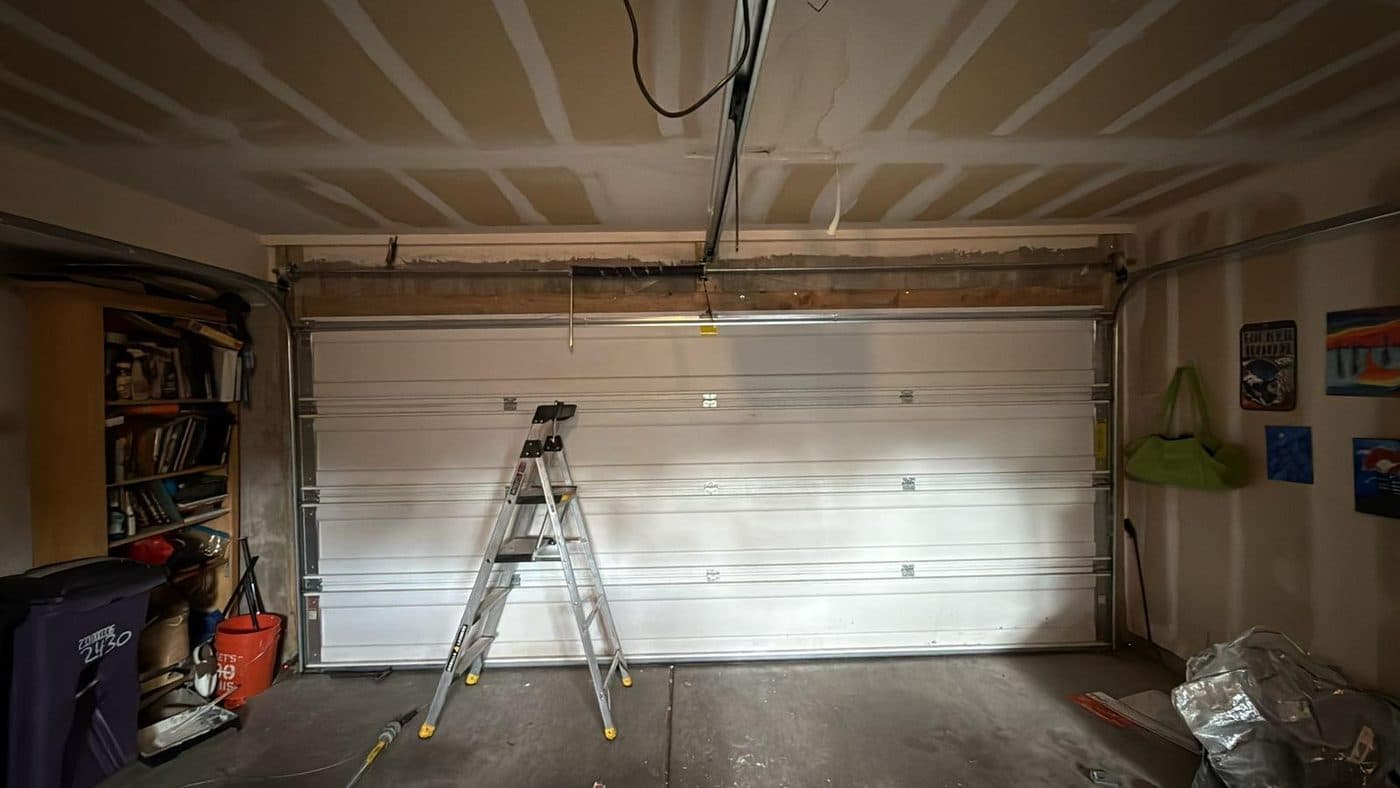 A step ladder standing in front of a white sectional garage door part-way through a job, tools on the floor.